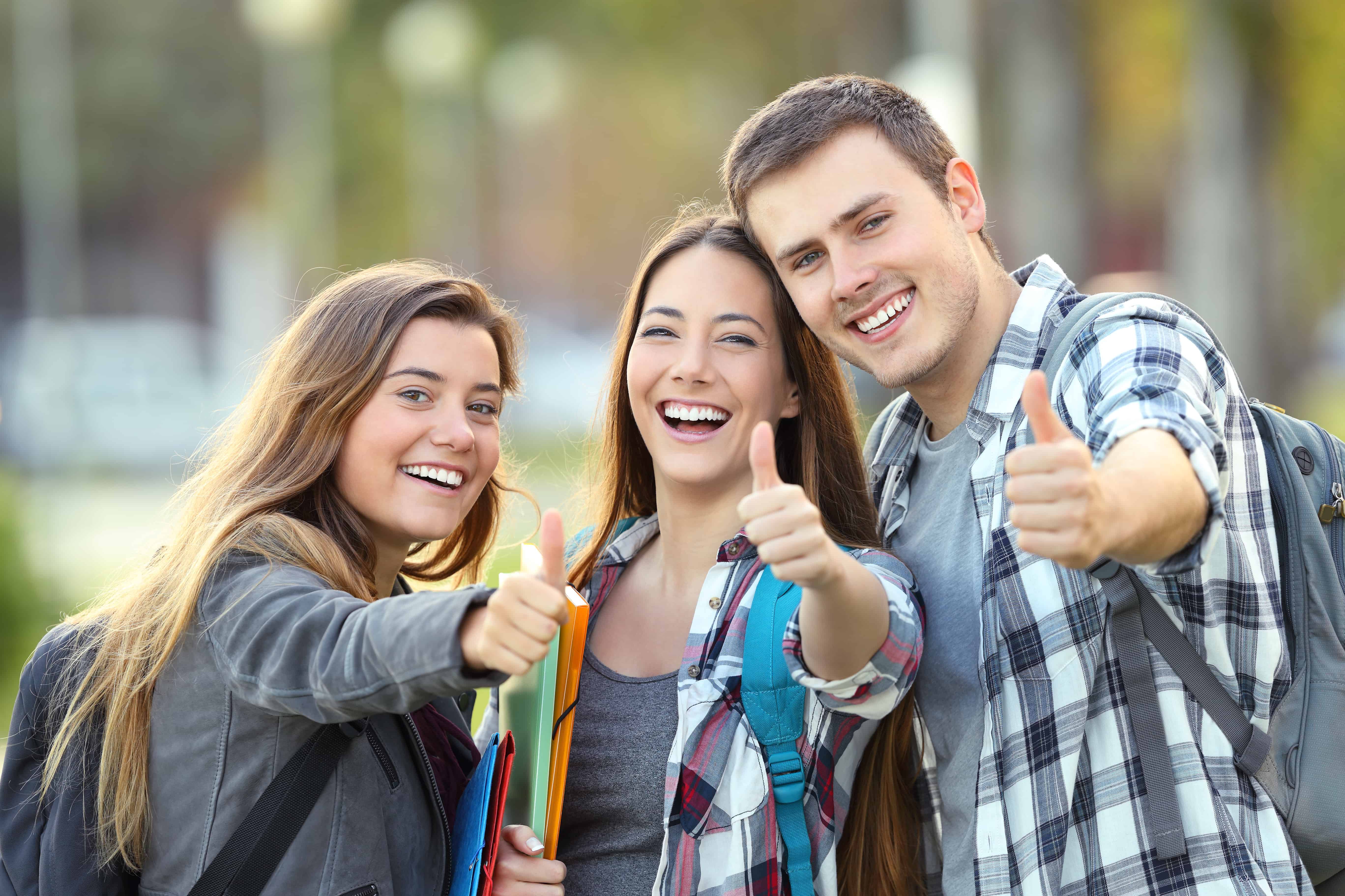 Three happy students looking at you with thumbs up in an university campus