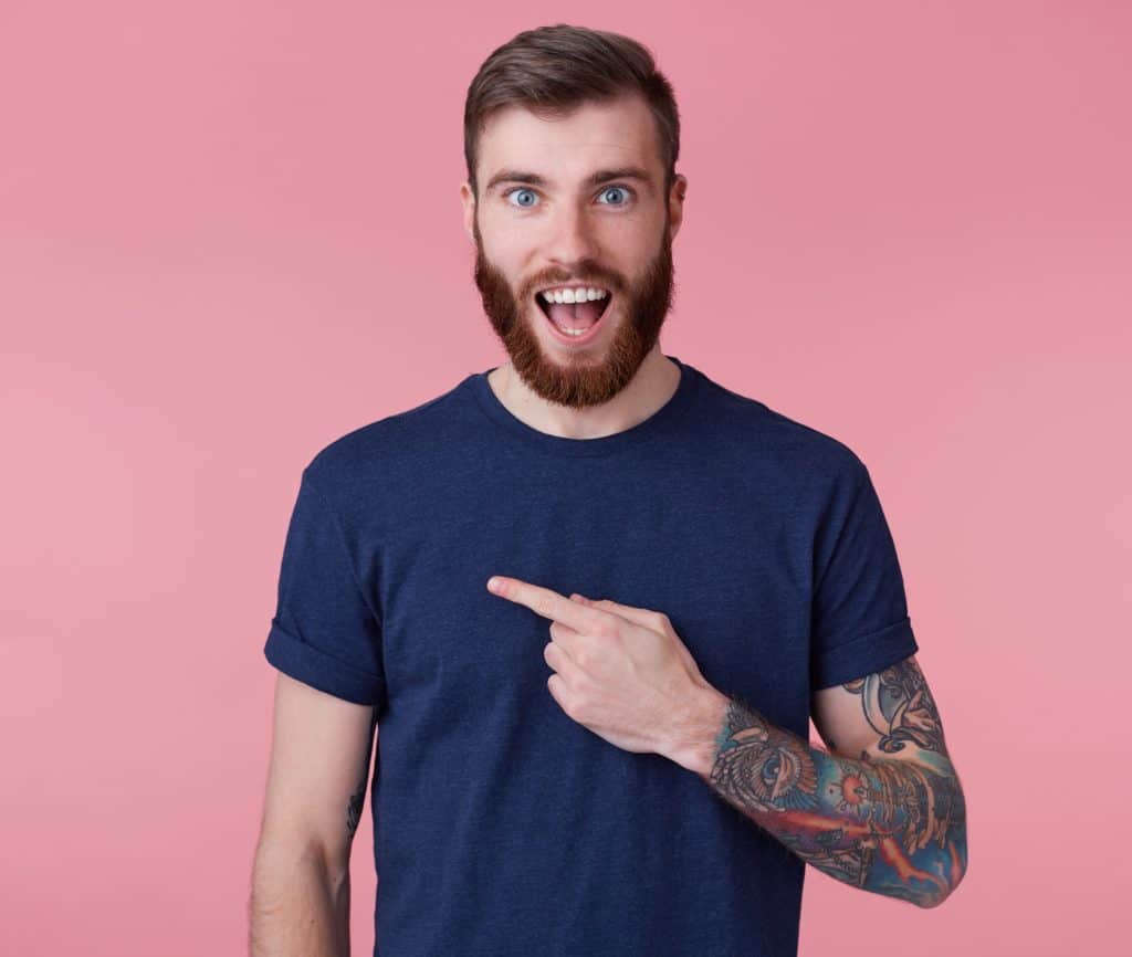 Young happy amazed attractive red-bearded young guy , wearing a blue t-shirt, with wide open mouth in surprise, pointing finger to copy space at the left side isolated over pink background.